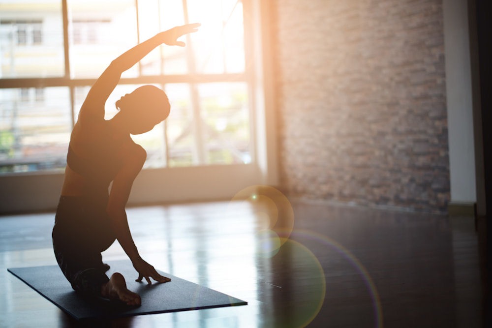 Silhouette of a woman stretching on a yoga mat in front of a large window with sunlight, symbolizing mindfulness during National Yoga Awareness Month.